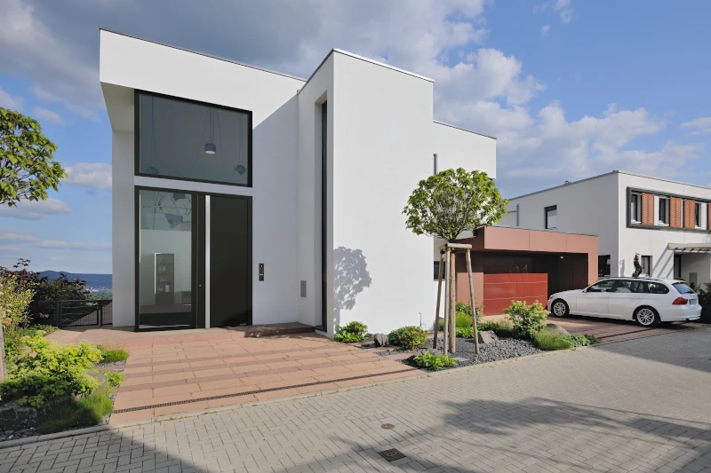 Ein kubischer Bau eines Hauses mit einer weißen Fassade und schwarzen Fenster sowie Türelementen. Der Himmel ist leicht bewölkt.
