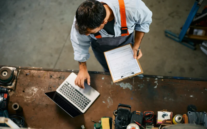 Handwerker steht mit Klemmbrett und Laptop in der Werkstatt. Ansicht von Oben.
