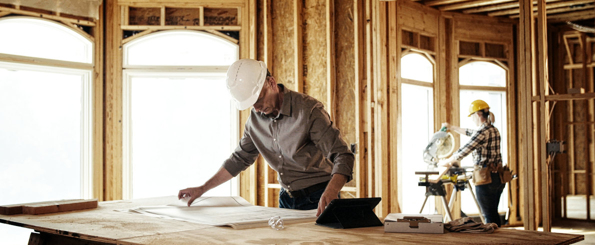 Zwei Bauarbeiter arbeiten in einem Gebäude mit Holzrahmen. Einer inspiziert Baupläne, der andere bedient eine elektrische Säge. Helle Fenster im Hintergrund.