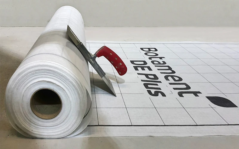 Das Bild zeigt eine aufgerollte Bahn mit der Aufschrift „Botament DE Plus“. Ein Cuttermesser mit rotem Griff liegt darauf, und der Untergrund ist mit einem Rastermuster versehen.