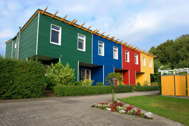 Vier Reihenhäuser, die nebeneinander stehen. Die Häuser haben Fassaden aus Holz, jedes in einer anderen Farbe: grün, blau, rot und gelb.