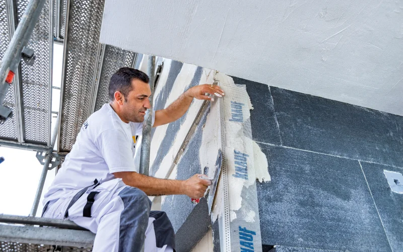 Handwerker sitzt auf Gerüst und verstreicht Putz auf einer Innenwand mit einer Kelle