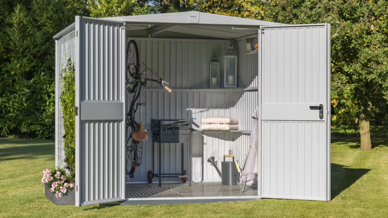 Ein offenes Gartenhaus in einem Garten mit Gartenzubehör, wie einem Fahrrad, Grill, Lampen und Gieskanne, befüllt.