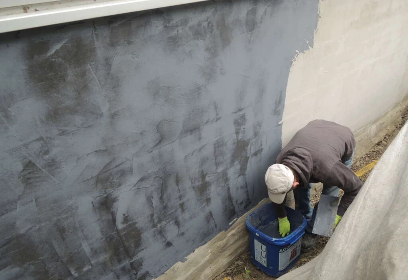 Ein man, der eine Fassade die mit mineralischer Bauwerksabdichtung bestreicht. Der Mann rührt in einem Eimer.