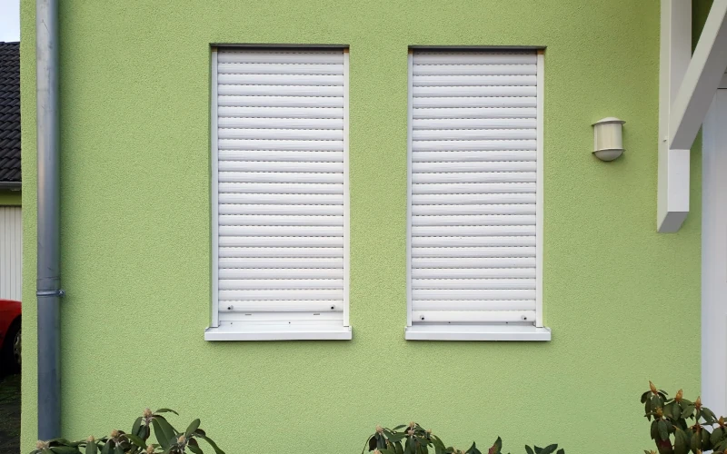 Zwei schmale Fenster in einem Haus mit grüner Fassadenfarbe haben einen weißen Außenrollladen