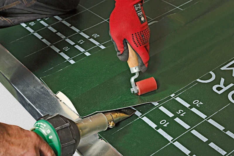 Handwerker mit roten Handschuhen verschweißt Dachfolie mit Heißluftgerät und Walze, präzise Arbeit.