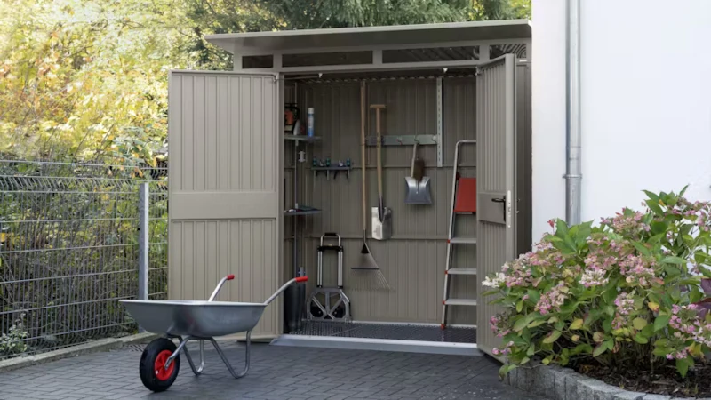 Ein kleines Gartenhäuschen welches offen steht und mit Gartenzubehör wie Schaufel und Leiter befüllt ist. Eine Schubkarre befindet sich vor dem Gartenhaus.