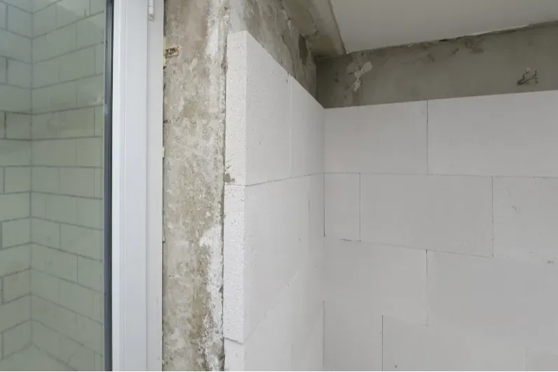Das Bild zeigt eine Wand mit unverputztem Beton links und neuen weißen Fliesen rechts. Oben ist die Wand unfertig. Links ist ein Glaselement sichtbar.