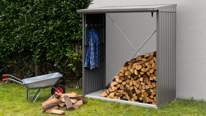 Ein Kaminholzregal im Garten wo Holz gelagert ist. Davor steht eine Schubkarre mit gespaltenem Kaminholz. In dem Kaminholzregal hängt noch eine Arbeitsjacke.