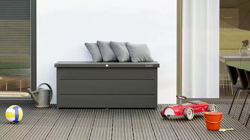 Eine Gartenbox auf einer Terrasse. Auf dem Terrassenboden liegt ein Ball, ein Speilzeugeimer und eine Schaufel, ein Bobbycar und eine Gießkanne.