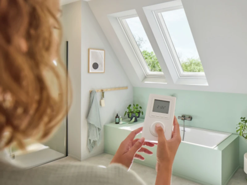 Frau misst mit digitalem Thermometer im Badezimmer die Temperatur. Im Hintergrund ist eine Badewanne und darüber zwei Heiz-Dachfenster.