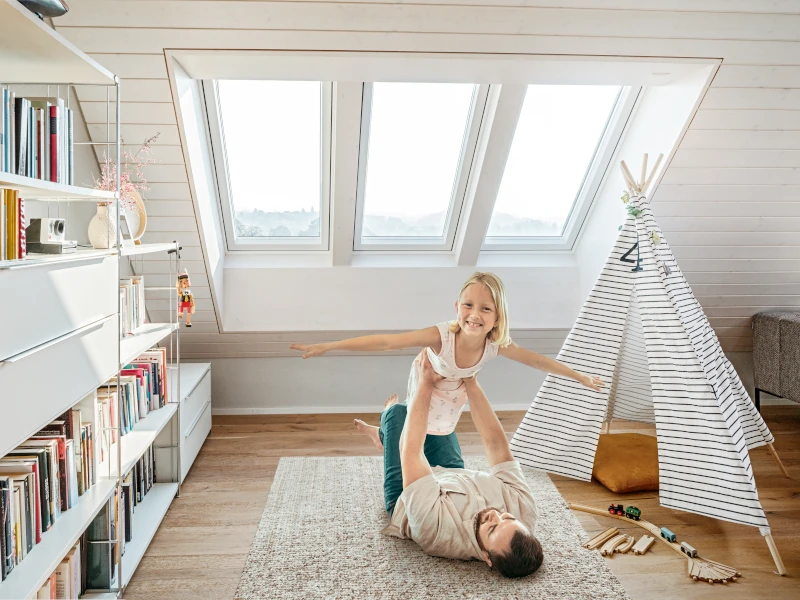 Mann liegt auf einem Teppich und hebt Mädchen hoch zum Spielen. Links steht ein Bücherregal und rechts ein Tipi-Zelt. Über den beiden Leuten sind 3 Heiz-Dachfenster eingebaut.