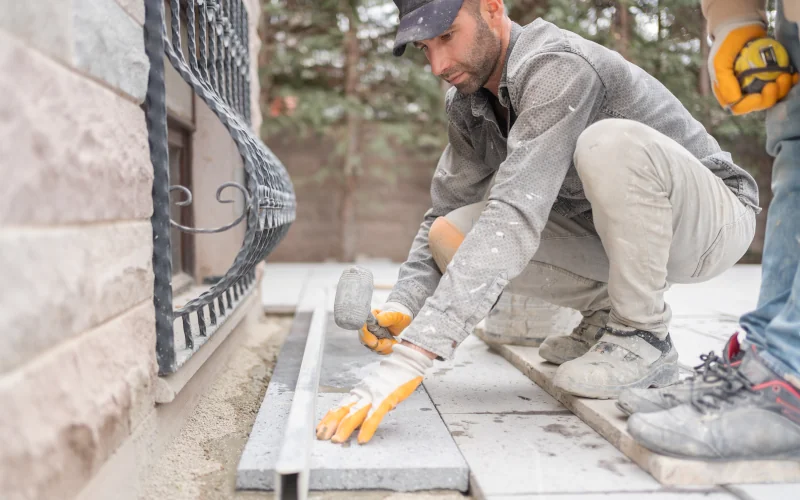 Handwerker knieend mit Hammer beim Verlegen von Terrassenplatten vor Gebäude