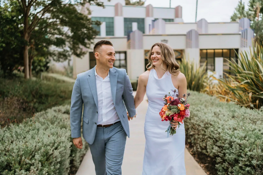 Couple standing on coastal path overlooking beach and cliffs at sunset, with calm ocean waters and soft evening light.