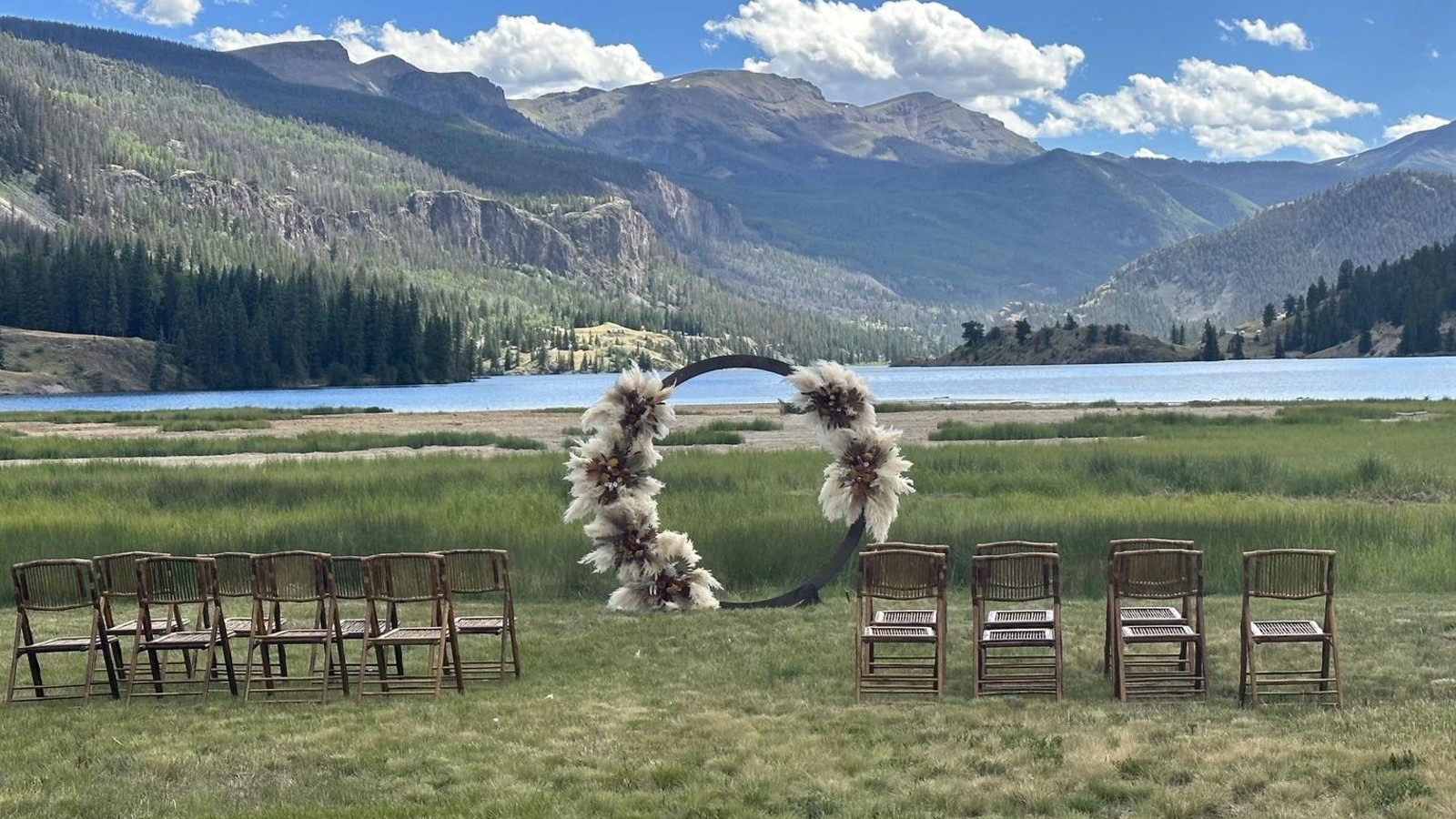 Outdoor wedding setup with floral arch and wooden chairs overlooking a mountain lake under blue sky with white clouds.