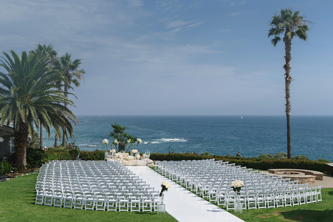 Oceanfront wedding venue with rows of white chairs, palm trees, and blue water backdrop under clear skies.