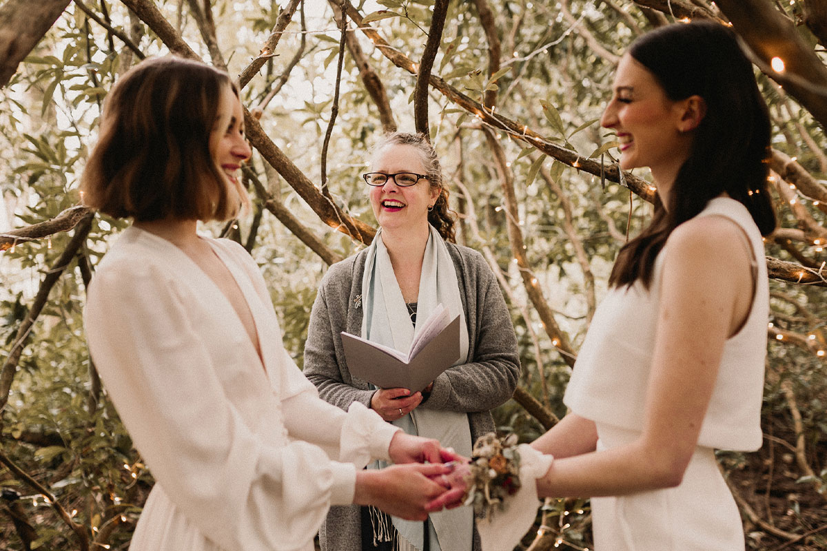 Two brides are holding hands in front of their wedding officiant, indicating they hired their officiant after researching wedding officiant costs.