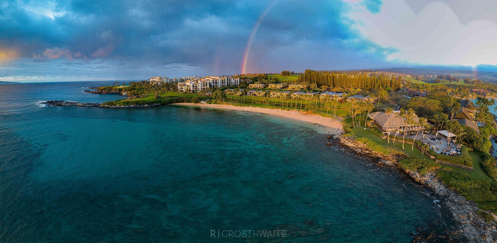 Aerial view of a tropical coastal resort with turquoise waters, sandy beach, and a rainbow arching over the landscape.