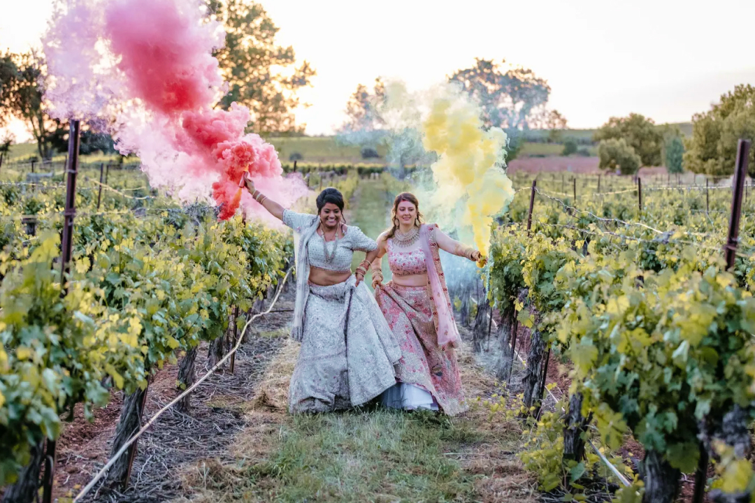 Two women in traditional lehengas walking through vineyard with colorful smoke bombs in pink and yellow at sunset.