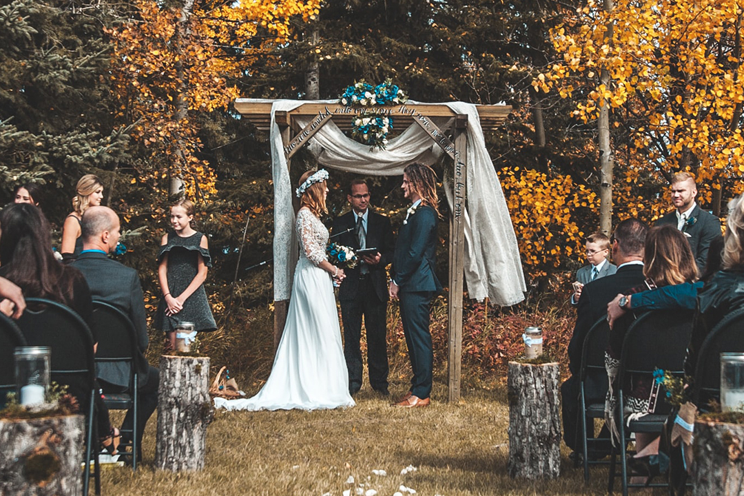 A bride and groom exchanging vows under a wooden arch outdoors in the fall.