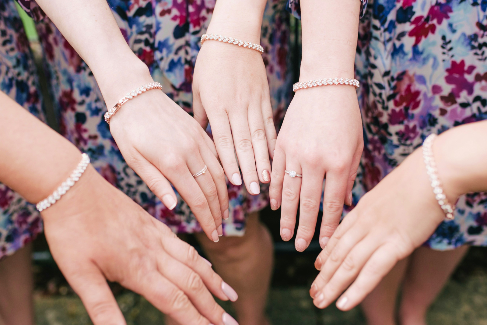 Four women's hands displaying elegant diamond tennis bracelets and rings, wearing floral dresses in purple and pink hues.