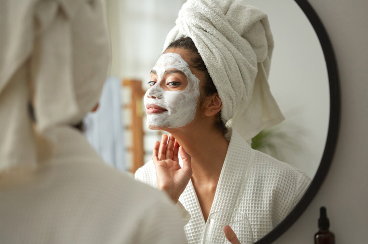 woman with hair towel and face mask looking in mirror.