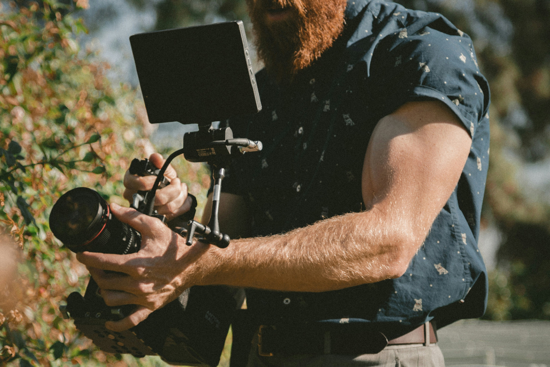 Person in blue shirt operating a camera with attached monitor outdoors among greenery.