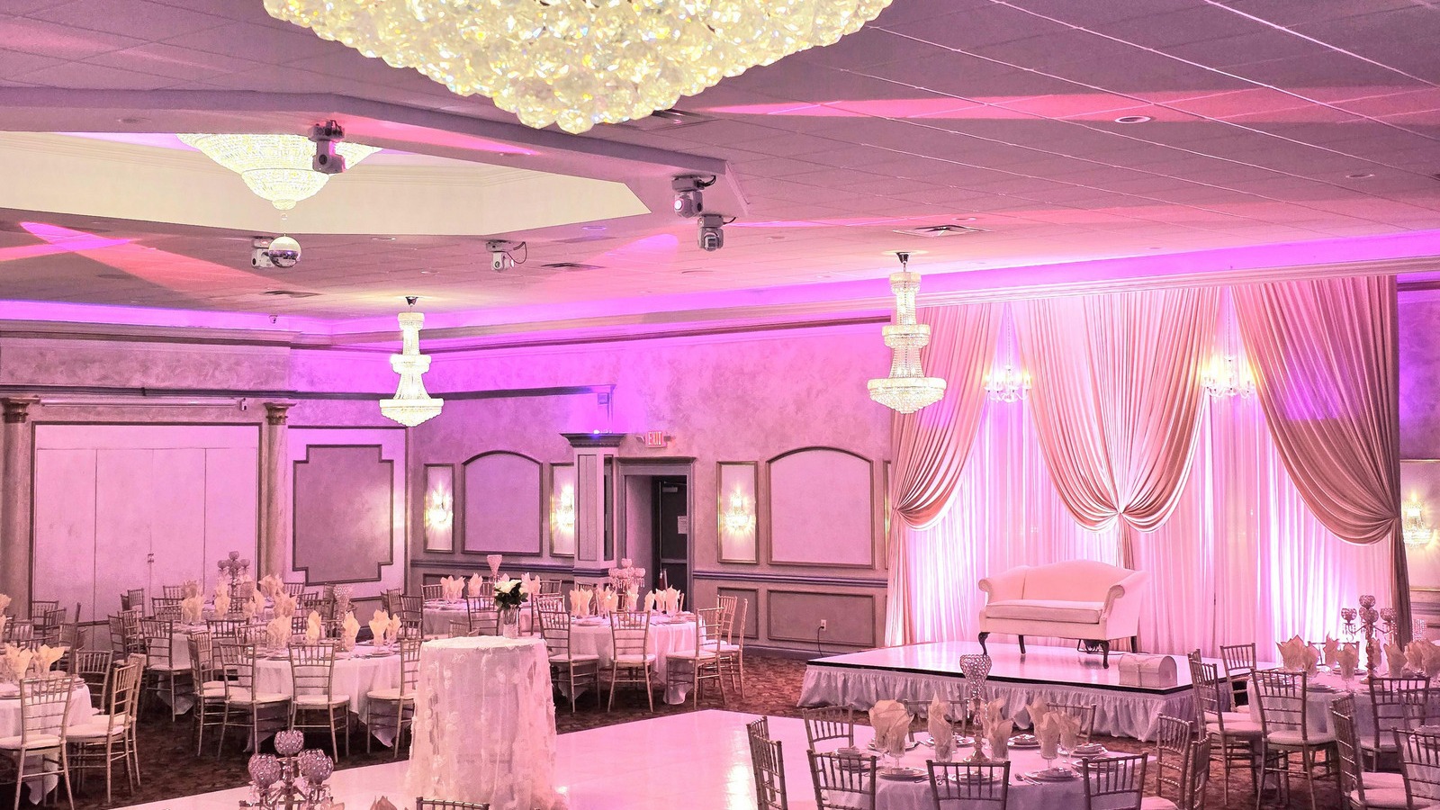 Elegant ballroom with crystal chandeliers, pink lighting, white drapery, and round tables set for a formal event.
