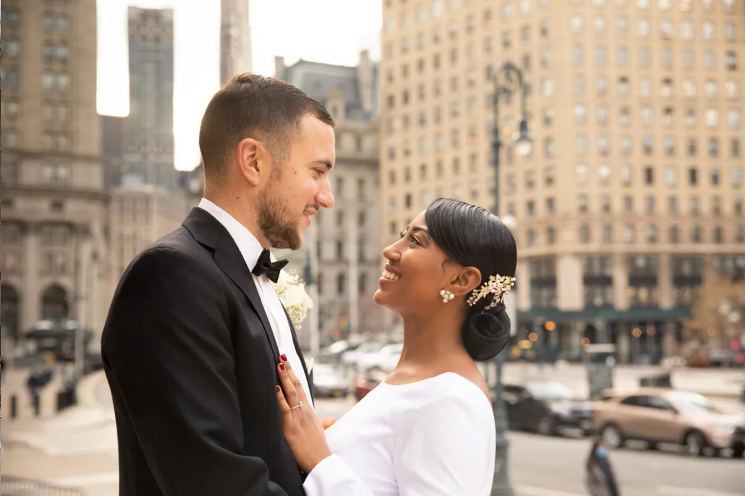 Couple in formal attire smiling at each other against New York City skyline with historic buildings in background.