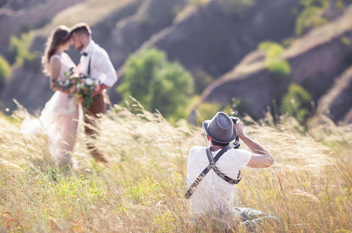 A wedding photographer crouches in a field to capture a shot of a husband and wife, potentially guiding them toward how to pose for wedding photos.