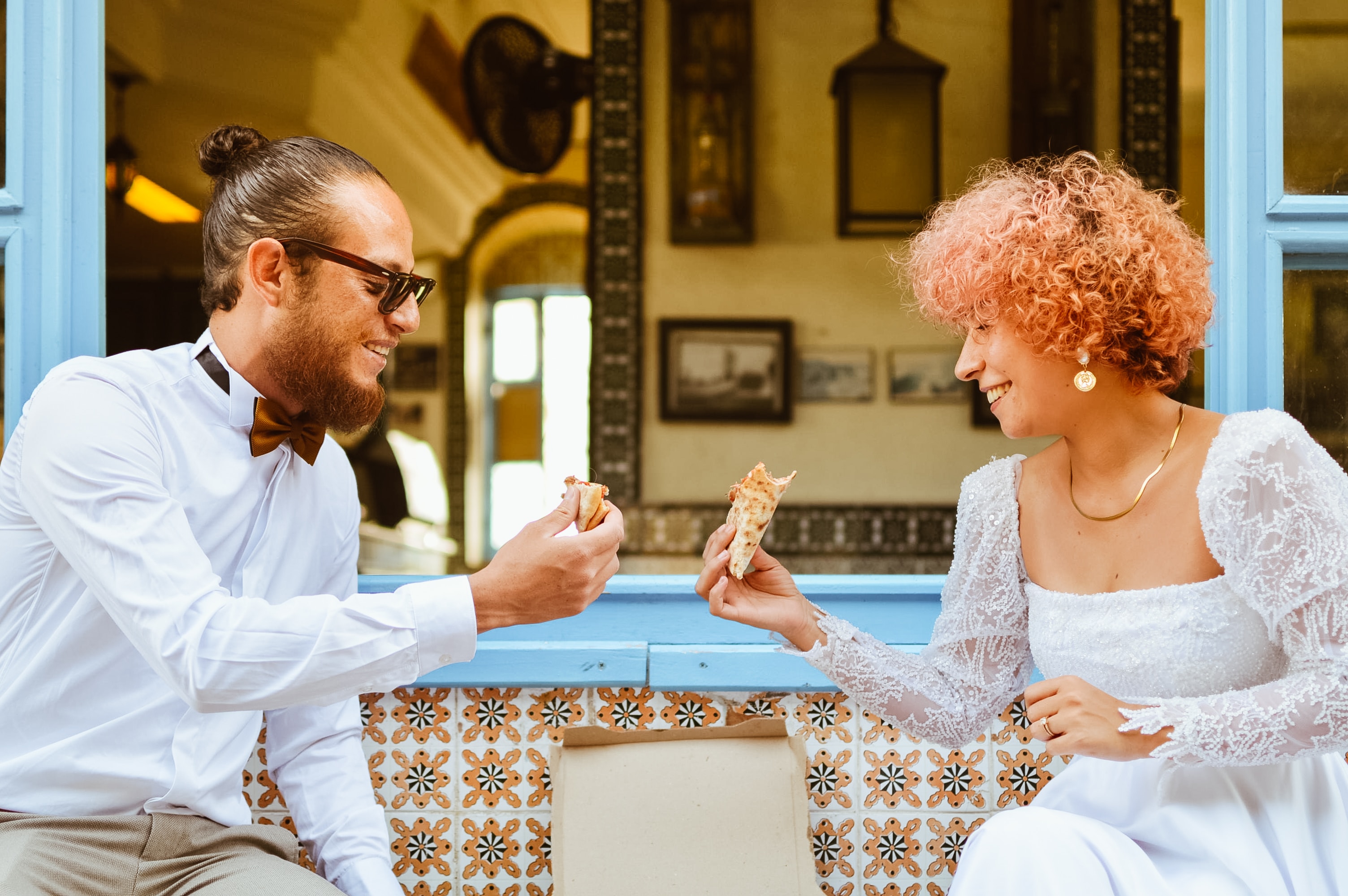 Couple in informal wedding attire toasting with slices of pizza.
