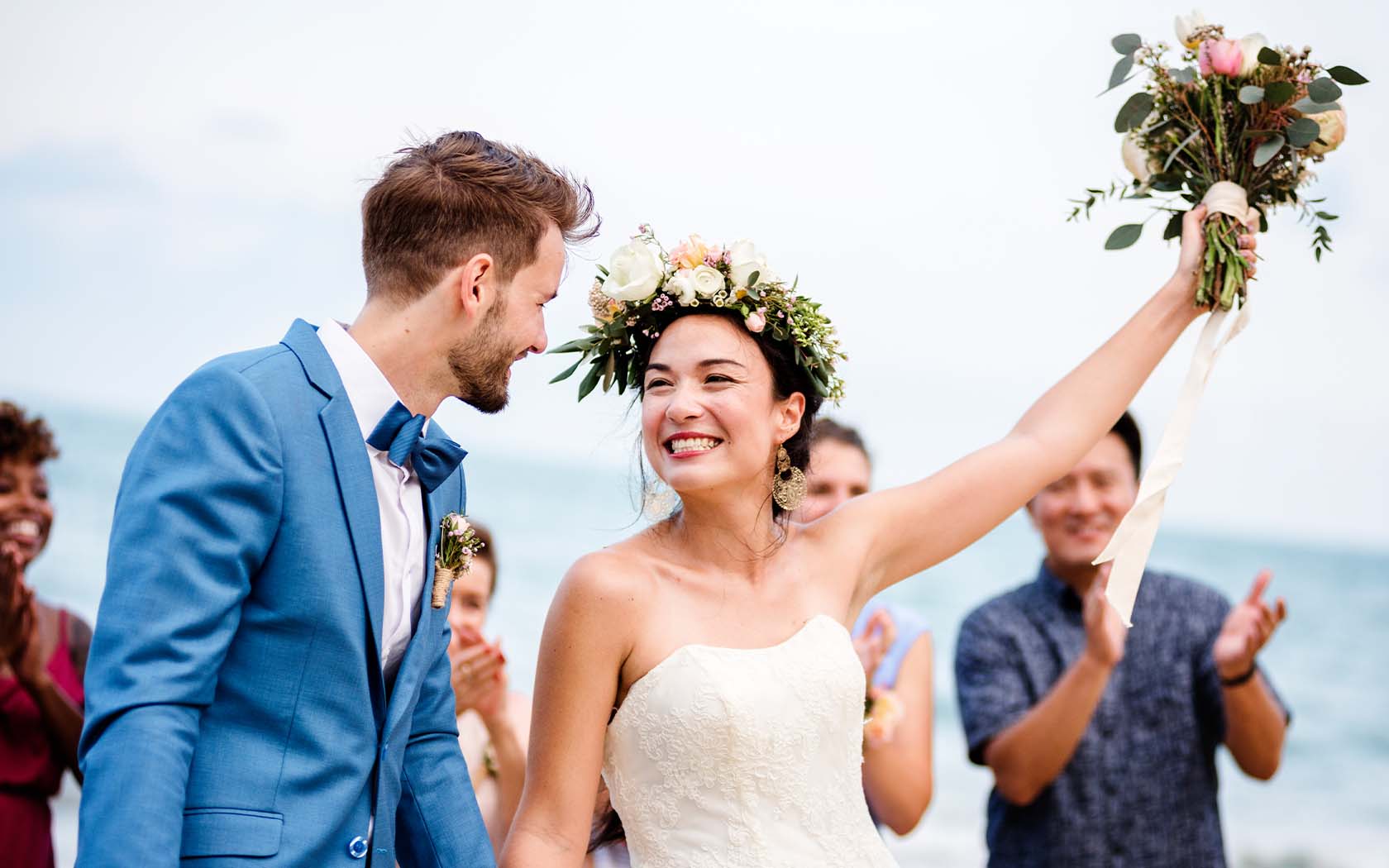 A bride is shown lifting her bouquet into the air and smiling at her new husband as wedding guests clap and cheer in the background.