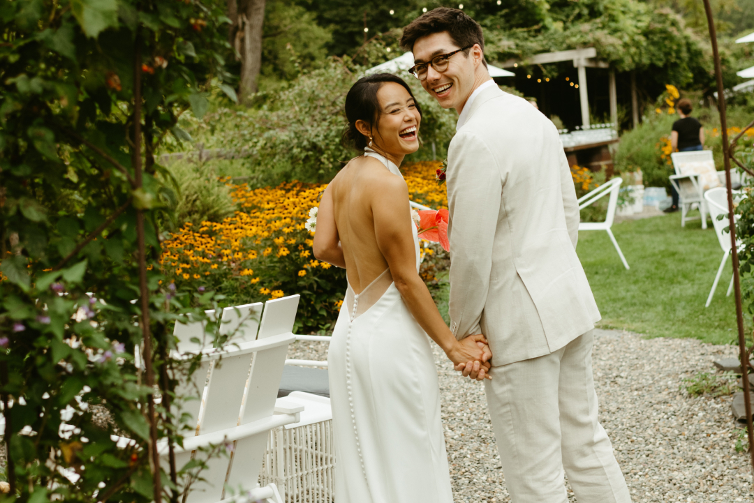 Happy couple in wedding attire holding hands in a garden with orange flowers and string lights among greenery.