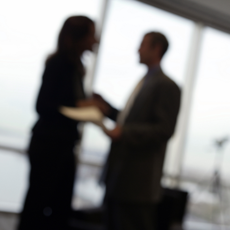 An image of a man and woman standing and speaking in a corporate office. They are in the background and out of focus, which makes the nature of their interaction unclear. They may be having an argument or a tense discussion.