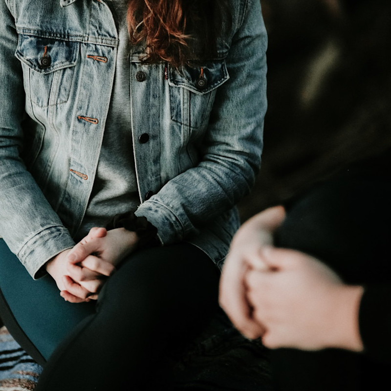 Woman in a denim jacket sitting with her hands clasped. In the foreground is a second figure in black; only their clasped hands are in shot, implying that a conversation is taking place between the two people.