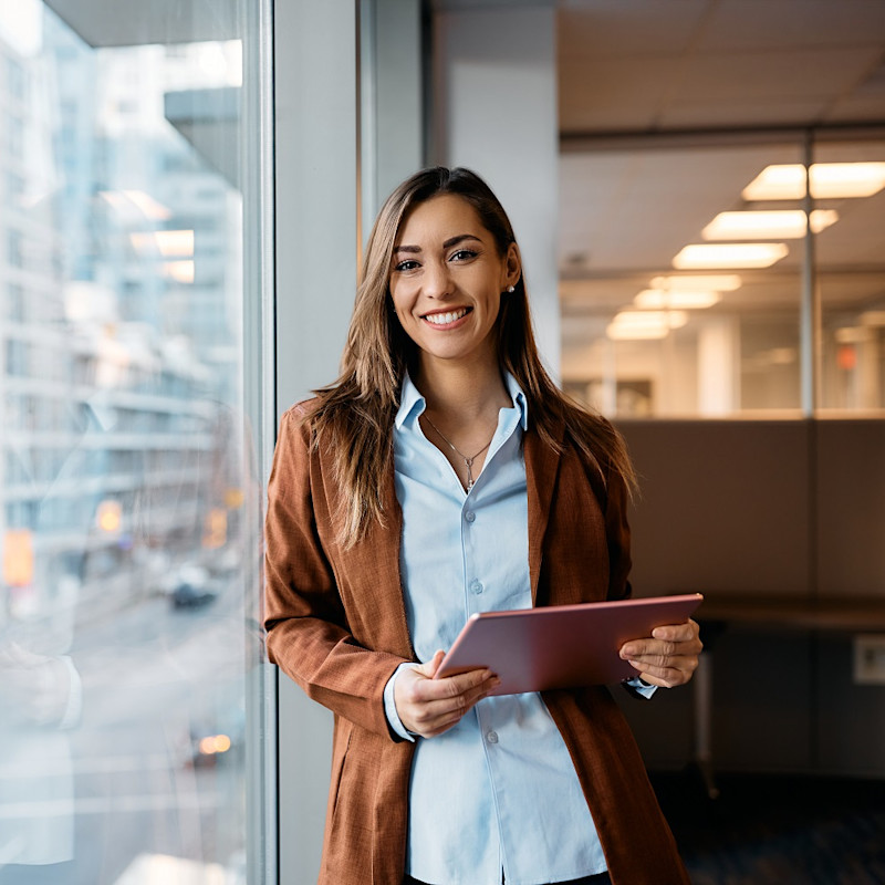 Lady leaning up against a glass window in an office holding an electronic device.