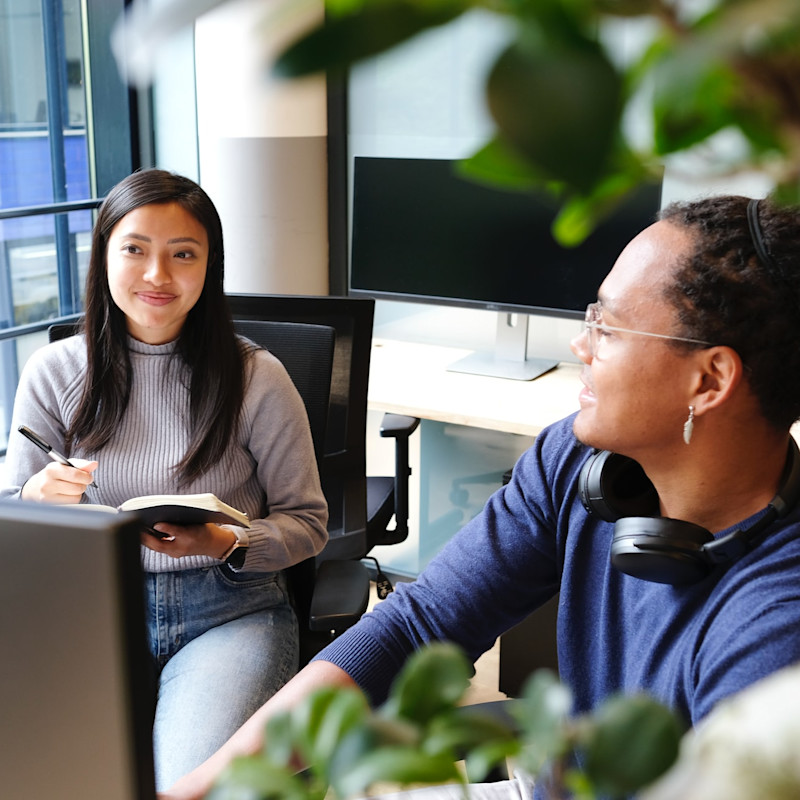 A man and woman sitting behind a desk in a corporate office environment. She is holding a notepad and taking notes.