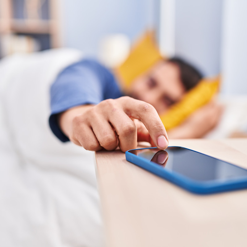 Man lying in bed, reaching his hand out to turn off his alarm or refuse a call coming through to the mobile phone lying on the sidetable next to him.