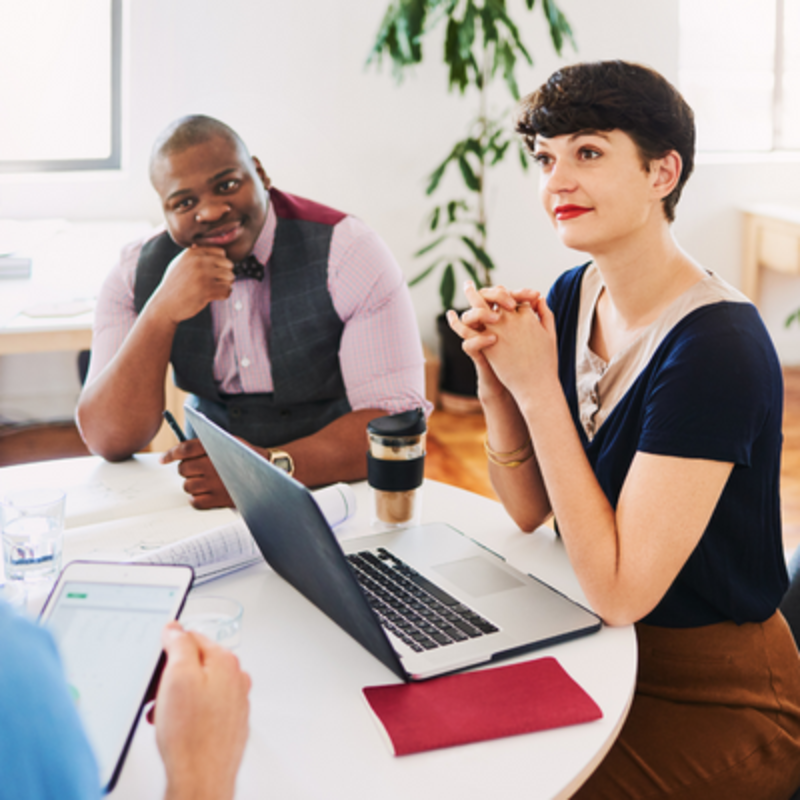 A woman is sitting in front of an open laptop. She is in an open-plan contemporary office with a cup of coffee next to her. Beside her sits a man leaning his chin on his hand. They are both looking at another man sitting in front of them holding a digital tablet.