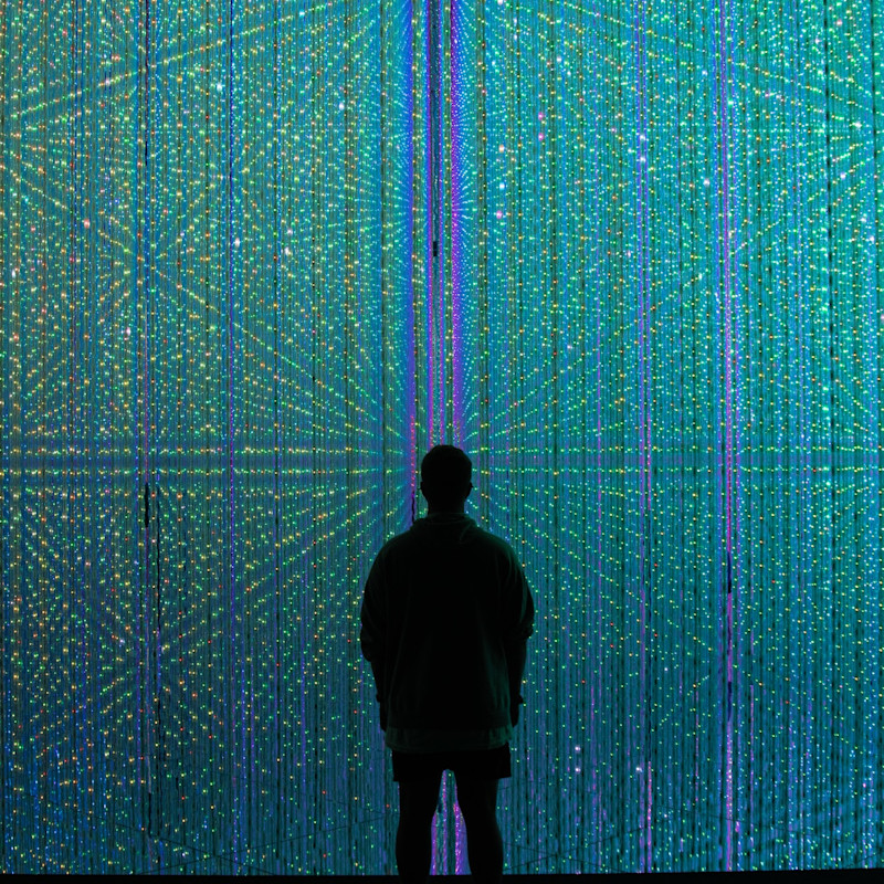 The silhouette of a man standing in front of a background of many small green and blue coloured lights.