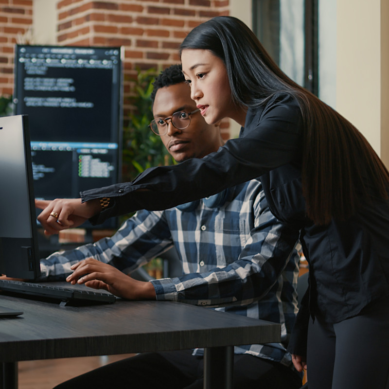 Two programmers or coworkers at an office desk working on a desktop computer.