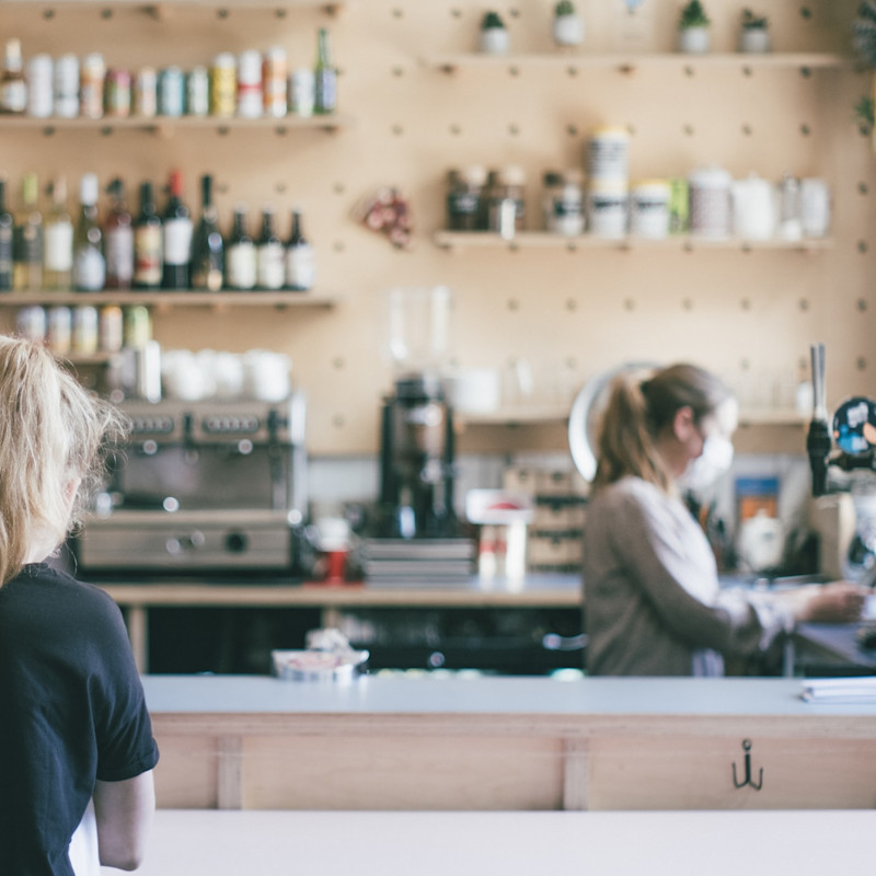 Two women standing at a cafe counter watching a barista or server at work.