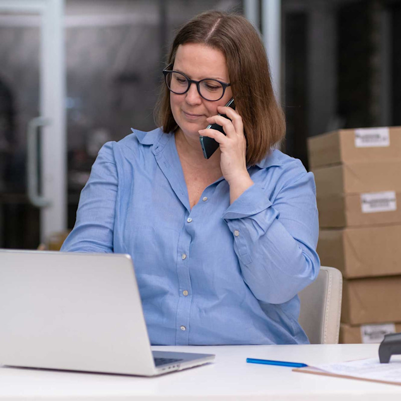 Business owner on mobile phone in front of laptop surrounded by cardboard boxes in a warehouse