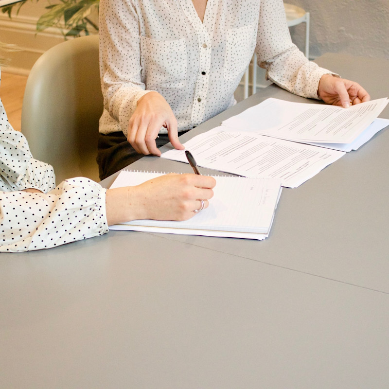 Two women, pictured from neck down, sitting at a desk and looking at or discussing documents or contracts laid out in front of them.