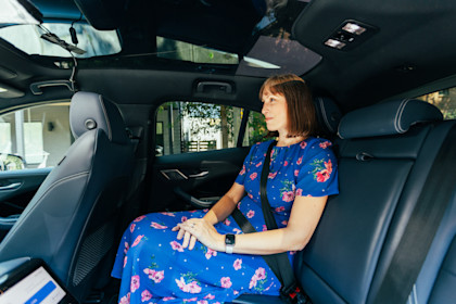 Suzy sits in the back seat of a Waymo vehicle, a Jaguar I-PACE, wearing a blue dress with pink floral patterns and a digital watch. She is looking out the window at a leafy neighborhood street in Atlanta, Georgia. The sunlight illuminates her face through the panoramic glass roof, and she is buckled in comfortably.