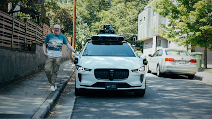 Robin Spinks stands on the sidewalk next to a Waymo vehicle, which is a white Jaguar I-PACE with several types of hardware sensors on the exterior. He is in a neighborhood in Atlanta, Georgia, with trees and houses in the background. He is wearing khaki trousers, a light blue and white shirt, blue glasses and is leaning on the vehicle with one hand resting just above one of the vehicle's radar sensors.