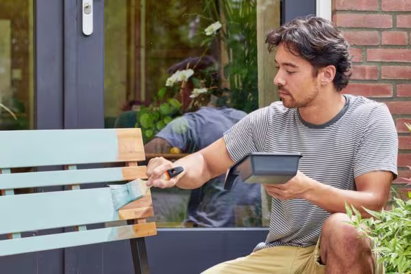 Persoon in gestreept shirt schildert een lichtblauwe houten bank in de tuin, met verfpot in de hand.