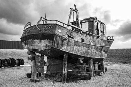 Photograph of stern of old rusting boat in shipyard propped up by large wooden blocks.