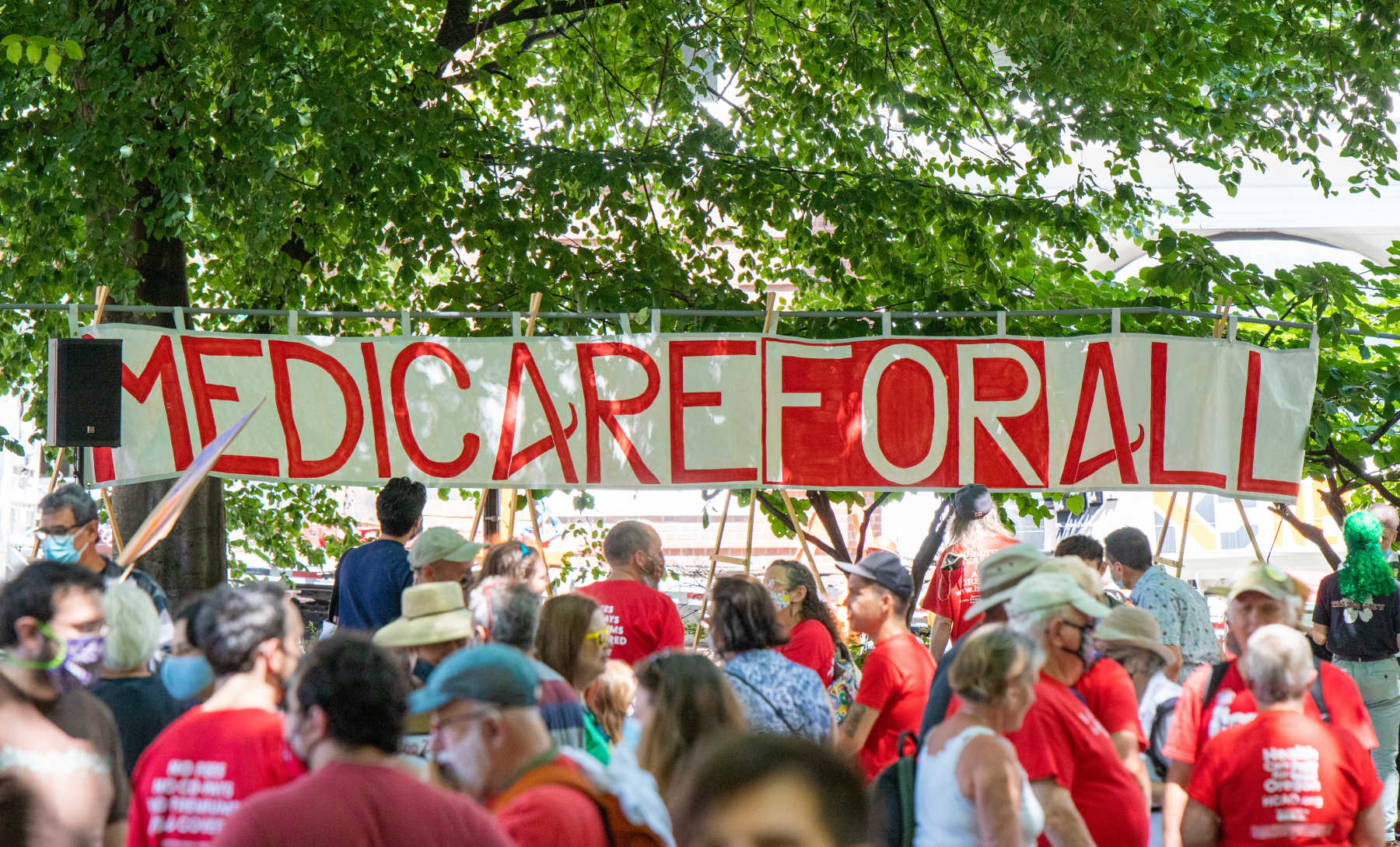 Many people wearing red shirts stand in front of a banner that says Medicare for All.
