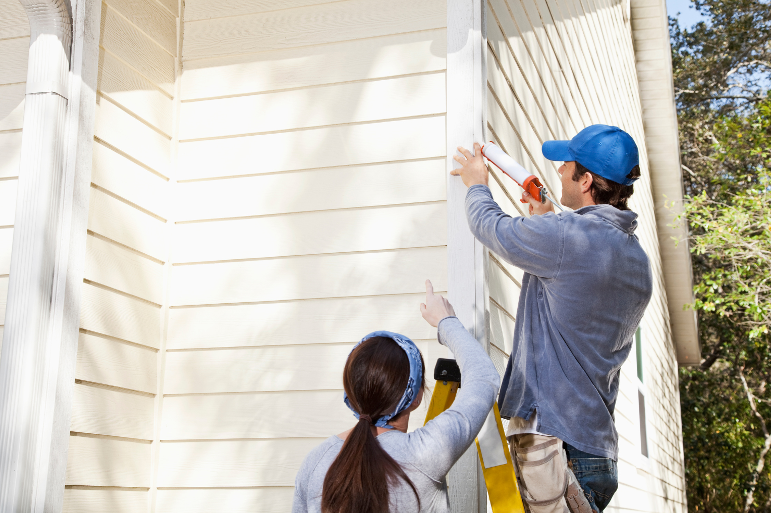 Professional contractor applying caulk to wood siding while homeowner observes the preventive maintenance work being performed on her house